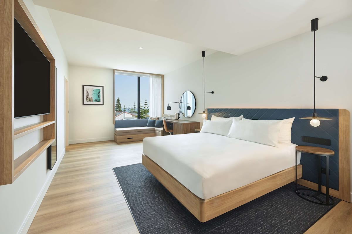 Habitación de hotel moderna con una cama grande, un escritorio, una silla, un televisor y una ventana con vistas a árboles.