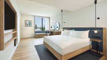A modern hotel room with a large bed, a desk, a chair, a TV, and a window with a view of trees.