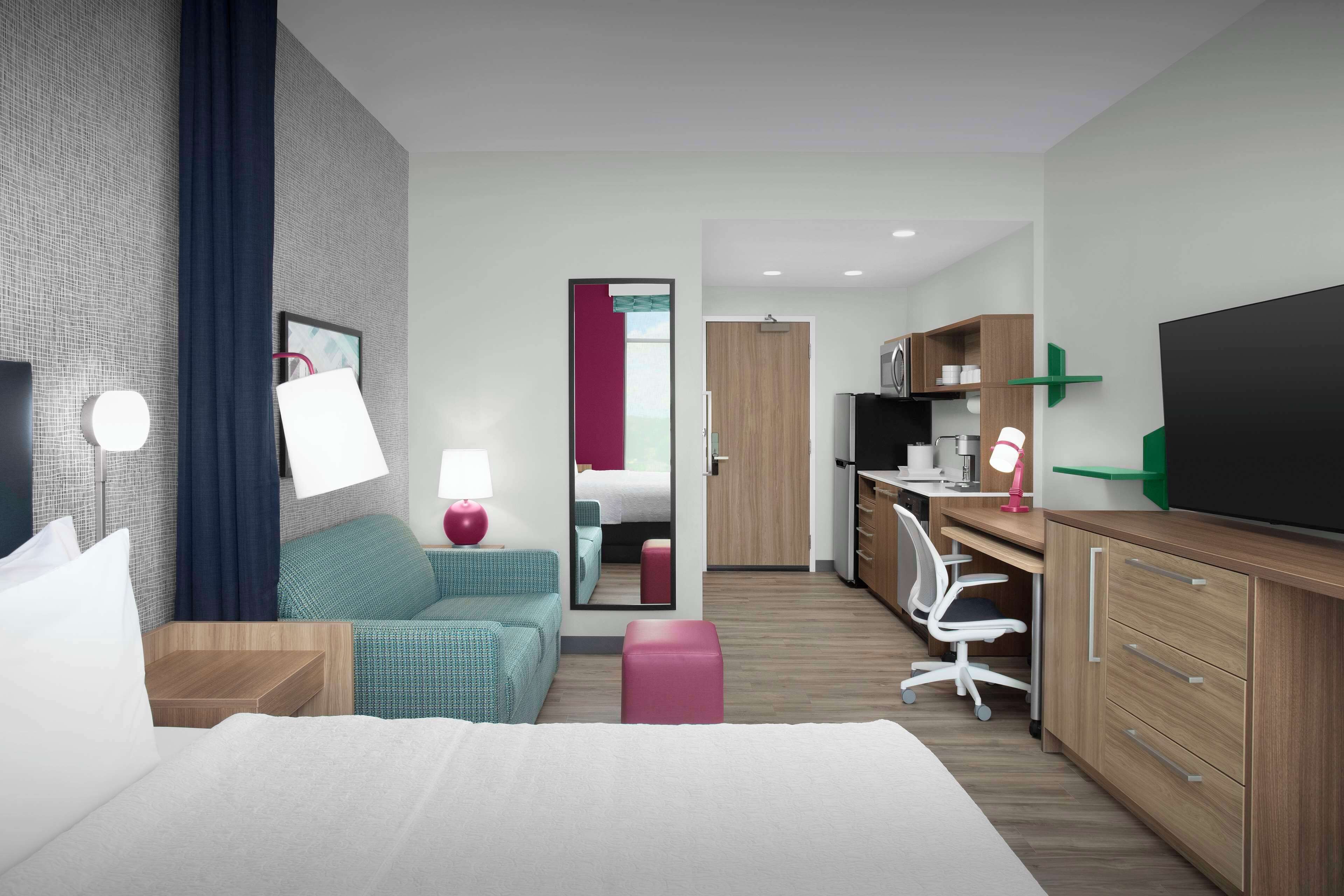 A modern hotel room with a bed, a desk, a chair, and a kitchenette.