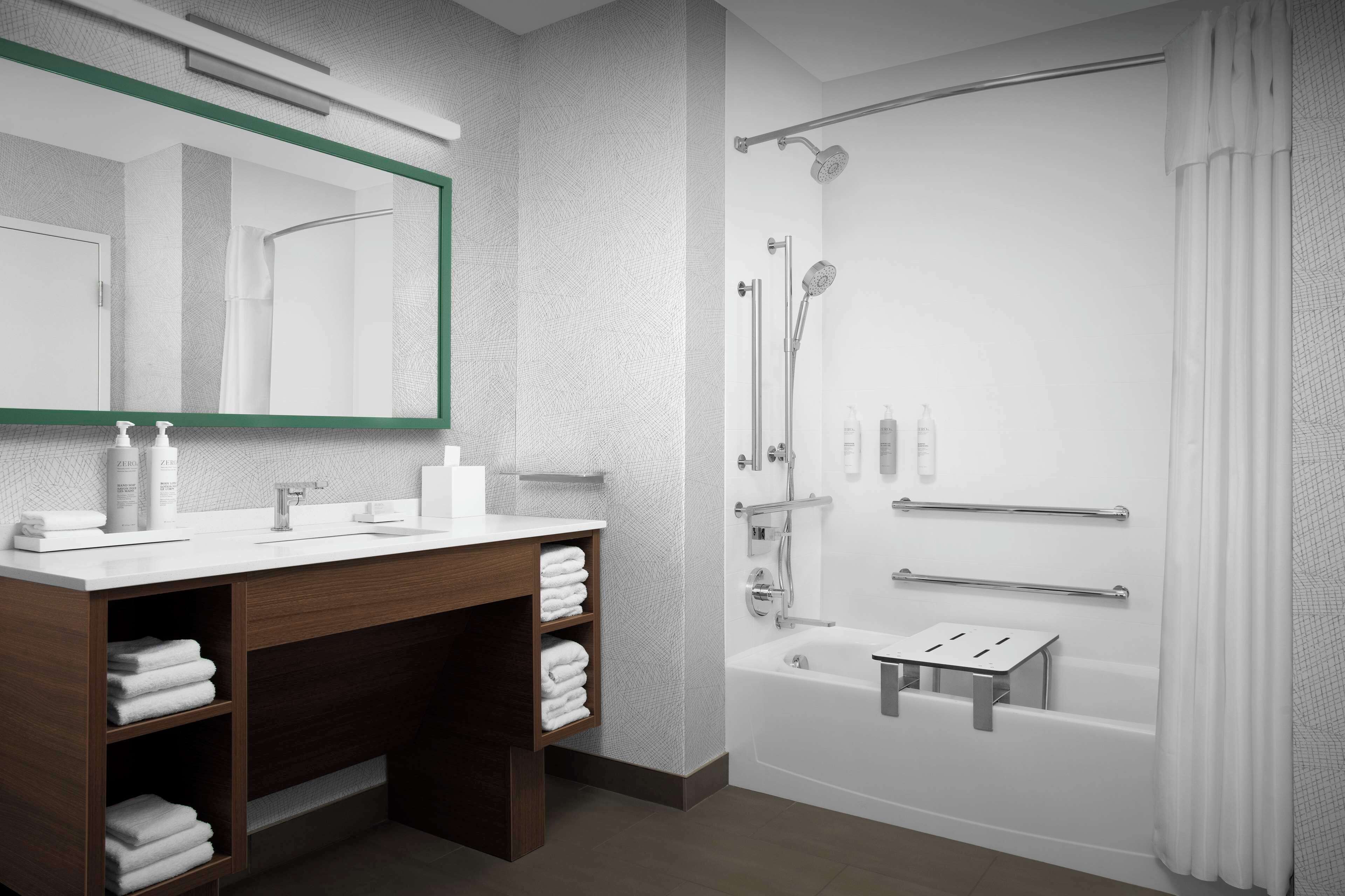 A modern bathroom with a wooden vanity, a large mirror, a shower with a handheld showerhead, and a small step stool.