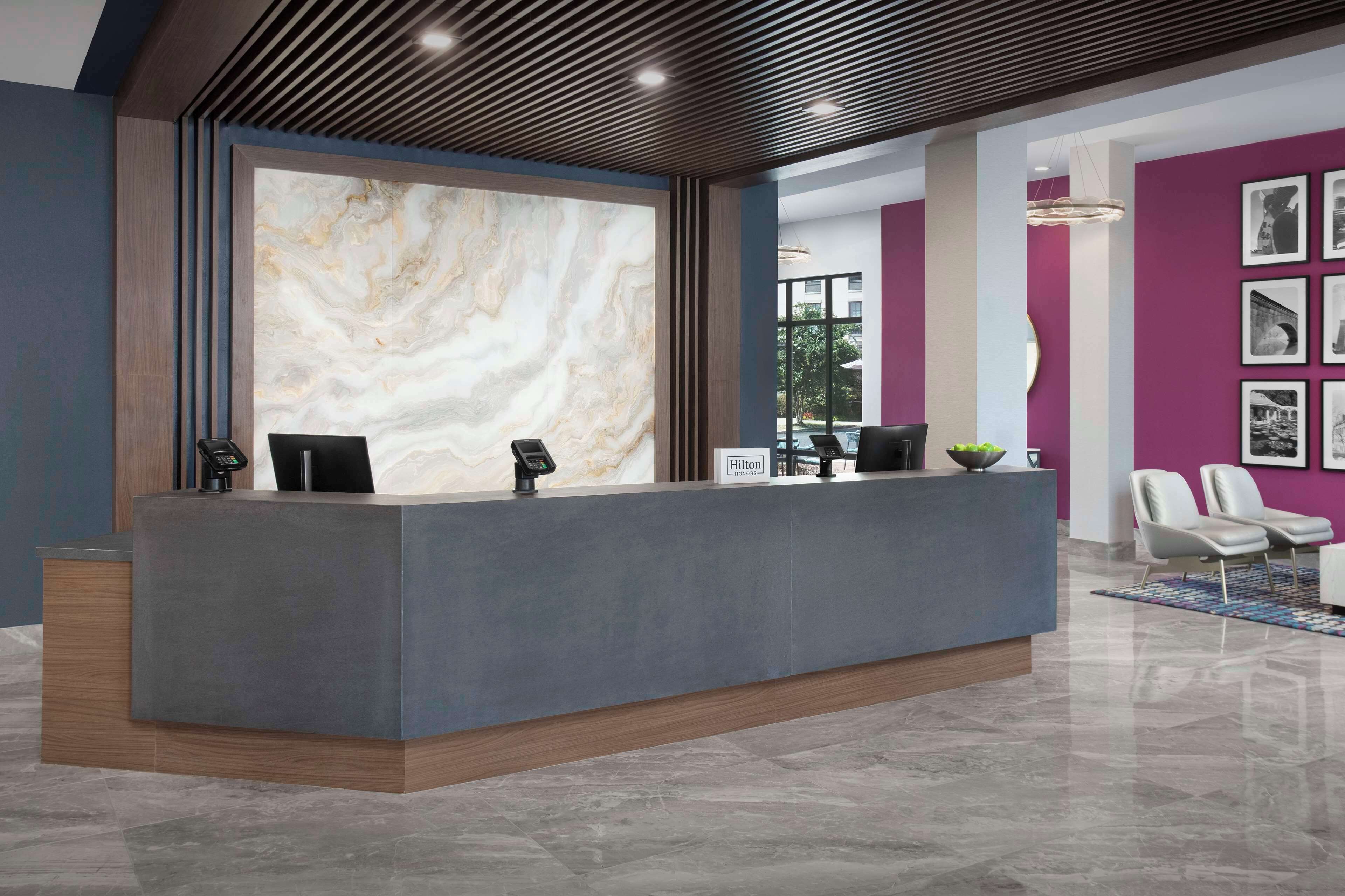 A modern hotel reception area with a marble wall, a grey reception desk, and a purple accent wall with framed pictures.