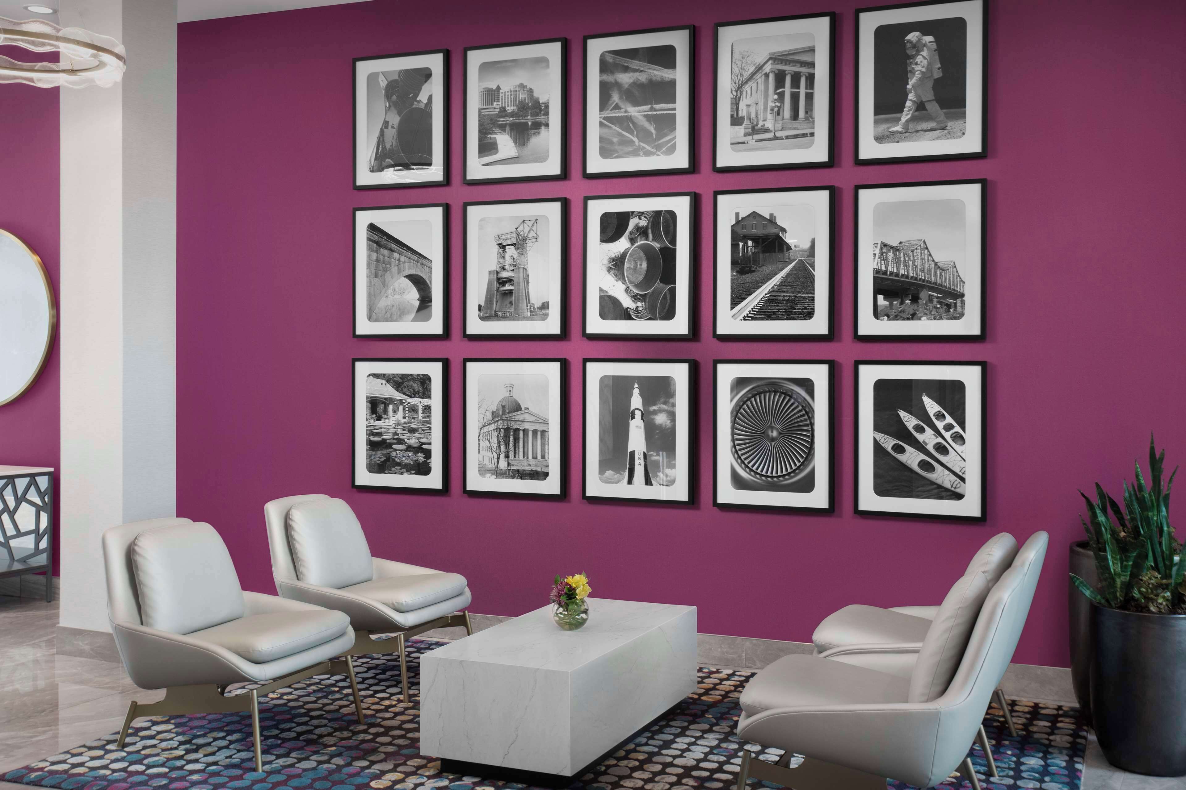 En moderne lobby med en lilla væg, sort-hvide billeder i rammer, to hvide læderstole, et centralt hvidt sofabord med en vase, og en tæppe med et mønster.