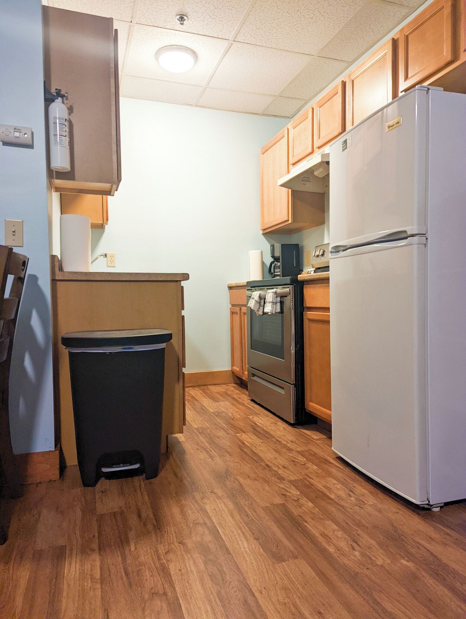 A kitchen with wooden cabinets, a stainless steel refrigerator, an oven, and a trash can.