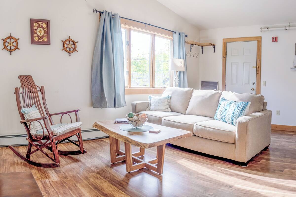 A cozy living room with a wooden rocking chair, a beige sofa with blue pillows, a wooden coffee table, and a large window with blue curtains.