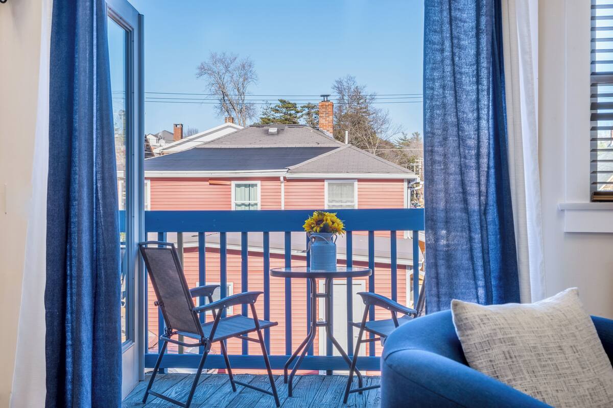 A balcony with a blue railing, a small table with a vase of yellow flowers, and two chairs.
