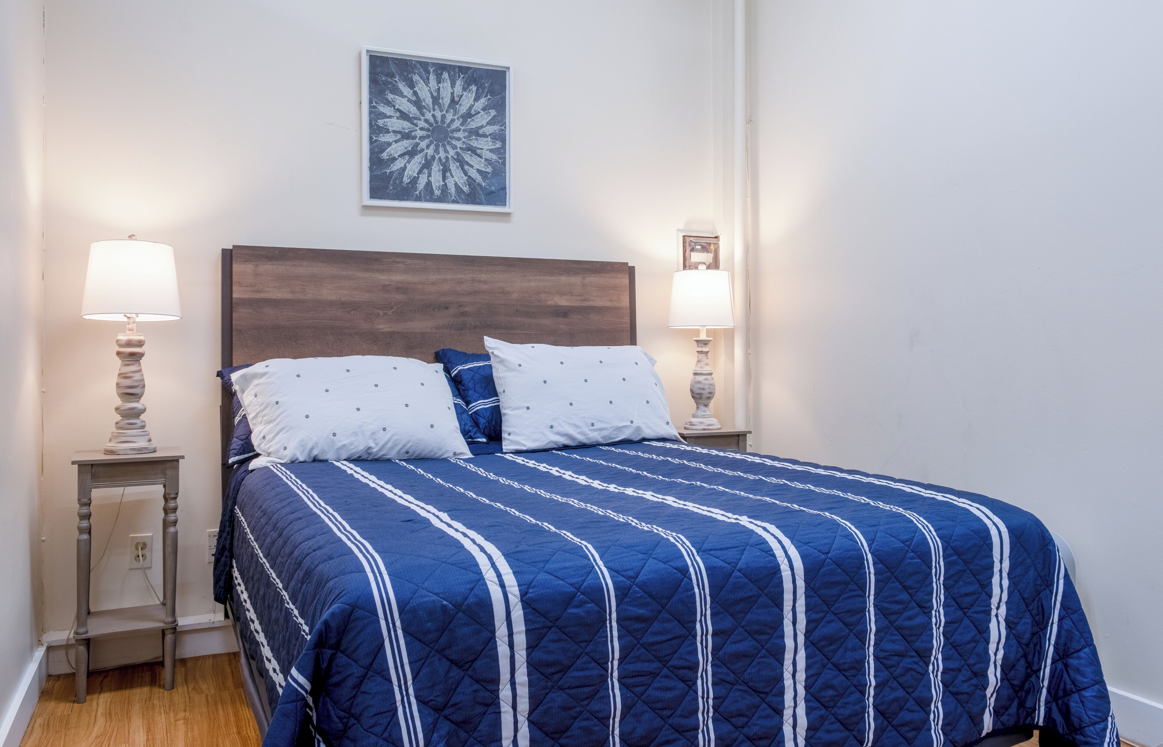 Un lit bien fait, recouvert d’une couette bleue et agrémenté d’oreillers blancs, est flanqué de deux tables de chevet avec des lampes. La pièce est caractérisée par un mur blanc sobre et une œuvre d’art florale accrochée au-dessus du lit.