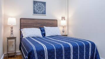 Un lit bien fait, recouvert d’une couette bleue et agrémenté d’oreillers blancs, est flanqué de deux tables de chevet avec des lampes. La pièce est caractérisée par un mur blanc sobre et une œuvre d’art florale accrochée au-dessus du lit.