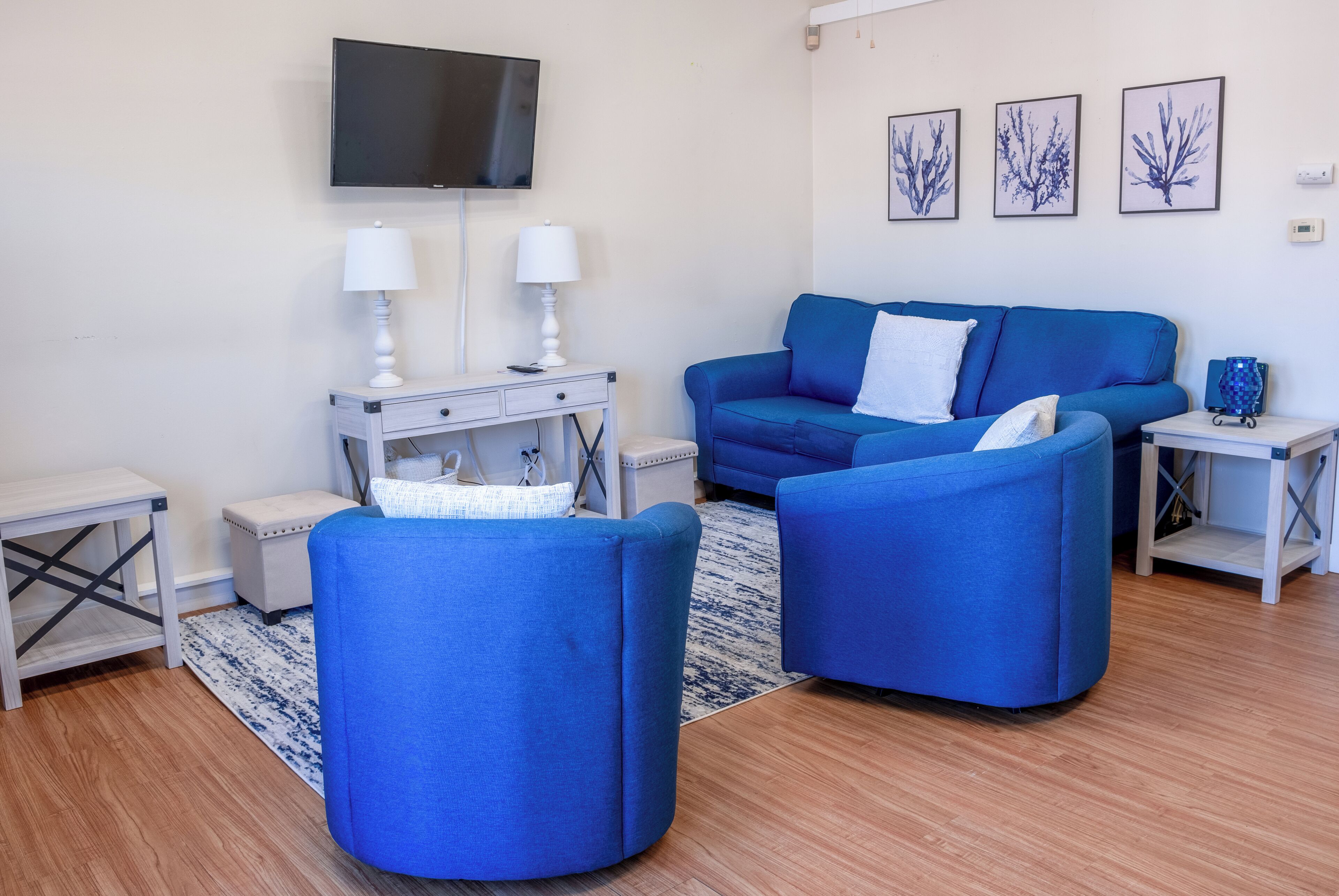 A modern living room with a blue sofa set, a flat-screen TV mounted on the wall, and a small white side table with a lamp.