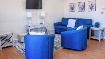 A modern living room with a blue sofa set, a flat-screen TV mounted on the wall, and a small white side table with a lamp.
