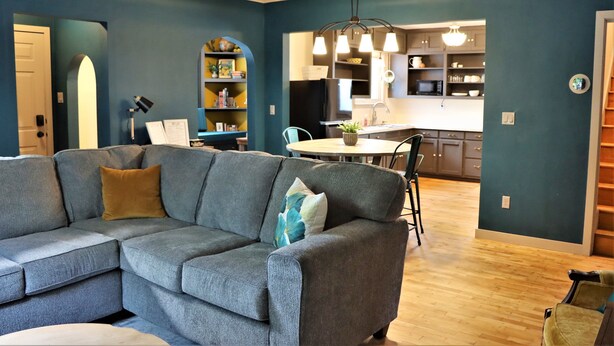 A living room with a grey sectional sofa, a dining table, and a kitchen in the background. - Columbia House. Unique, Comfortable Cape Cod Home. Pet friendly. (Green Bay)
