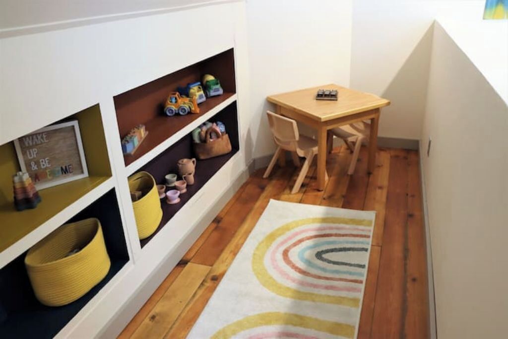 Een kamer met houten vloer, een houten tafel, een stoel en planken met speelgoed en opbergmanden.