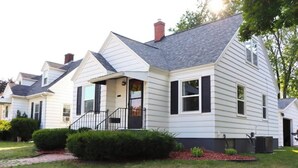 A single-story house with a front porch, a chimney, and a small front yard with a pathway and bushes. - Columbia House. Unique, Comfortable Cape Cod Home. Pet friendly. (Green Bay)