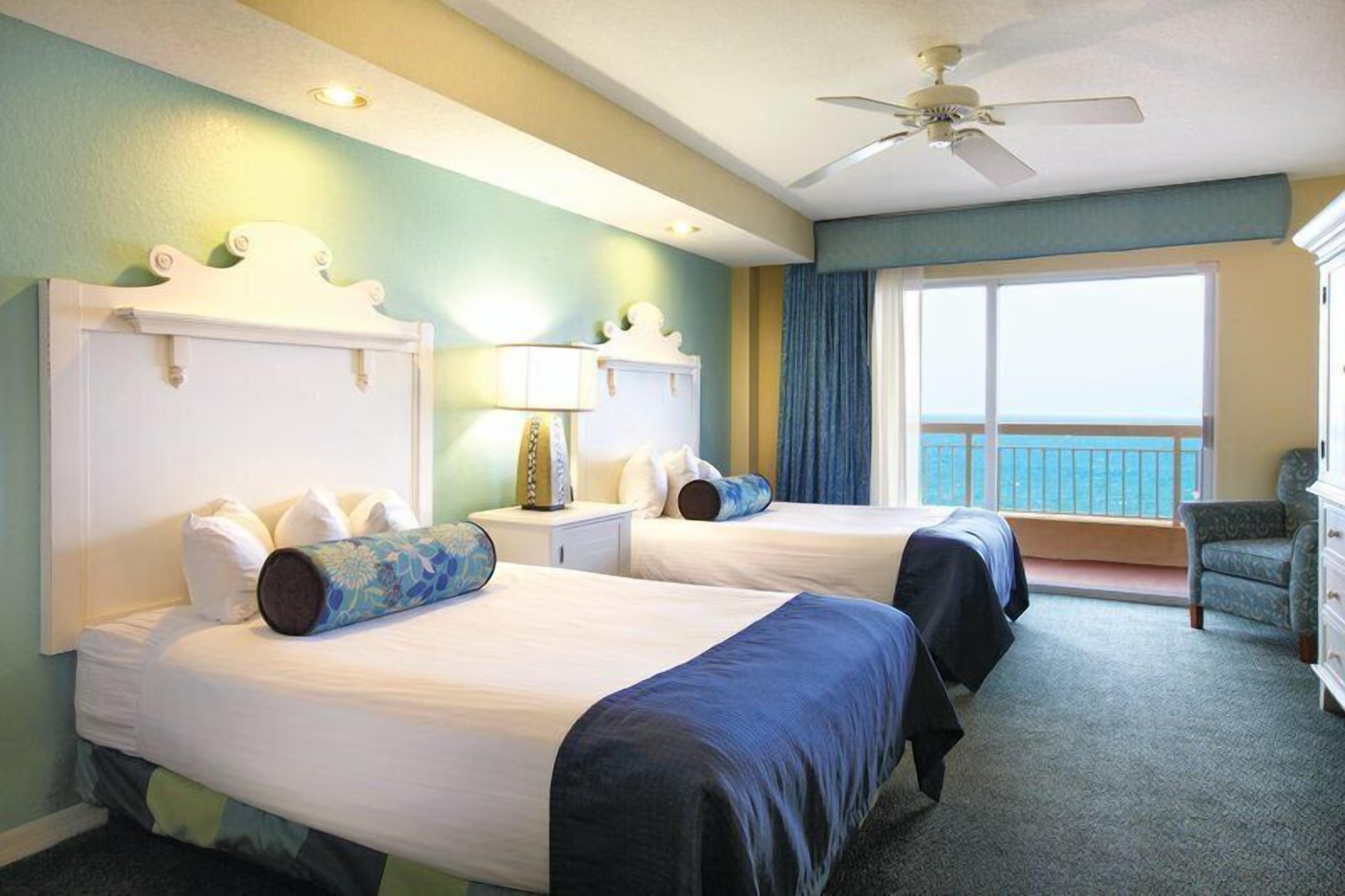 A hotel room with two beds, a balcony, a ceiling fan, and a view of the ocean.