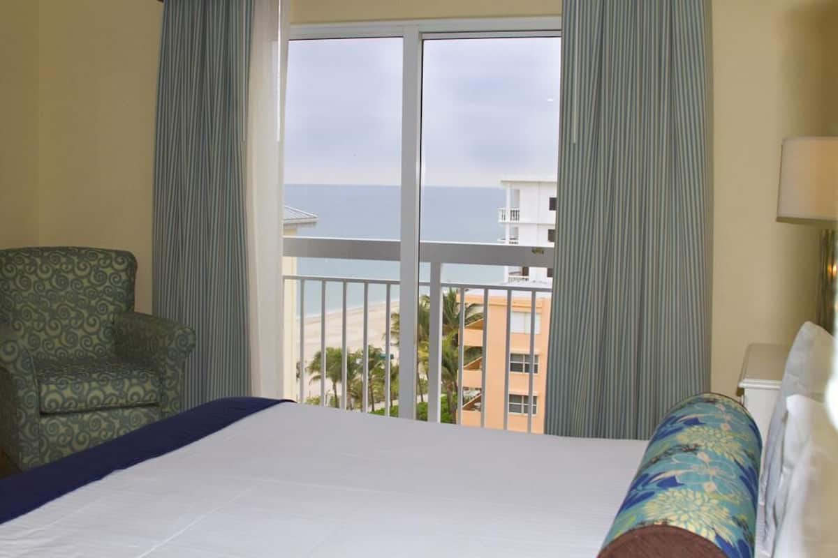 Ein Hotelzimmer mit Bett, Sessel, Balkon mit Blick auf das Meer und Gebäude sowie einer Lampe.