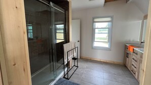 A bathroom with a glass shower door, a towel rack, a window, and wooden cabinets. - Four Season Waterfront Cottage - Private Escape | 3BR, 2BA | Howe Island (Gananoque)
