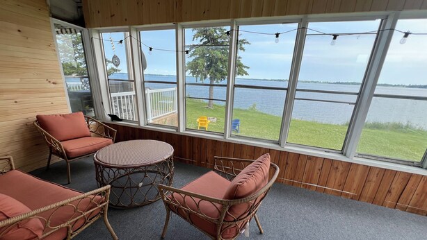 A covered patio with wicker furniture, large windows overlooking a lake, and a ceiling fan. - Four Season Waterfront Cottage - Private Escape | 3BR, 2BA | Howe Island (Gananoque)