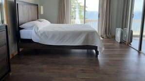 A bedroom with a bed, wooden floor, curtains, and a view of the outdoors. - Four Season Waterfront Cottage - Private Escape | 3BR, 2BA | Howe Island (Gananoque)