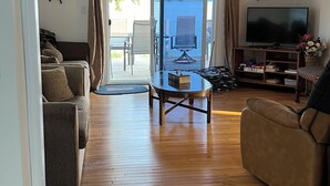 A spacious living room with a wooden floor, a ceiling fan, a television, and a view of the outdoors through a glass door. - Four Season Waterfront Cottage - Private Escape | 3BR, 2BA | Howe Island (Gananoque)