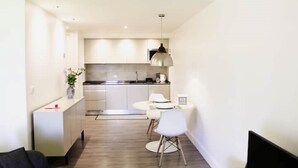 Un salon moderne et minimaliste, comprenant un canapé, une table à manger avec des chaises et un coin cuisine avec des placards et un luminaire suspendu.