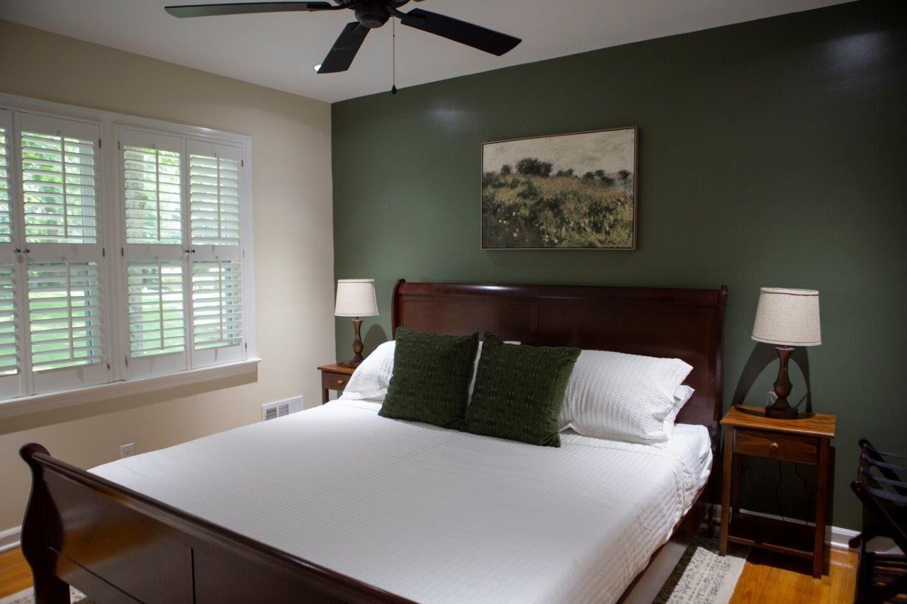A bedroom with a large bed, two bedside tables with lamps, a ceiling fan, a window with blinds, and a painting on the wall.