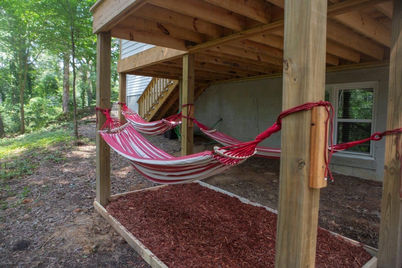 A wooden deck with a hammock suspended between two posts, surrounded by mulch and trees.