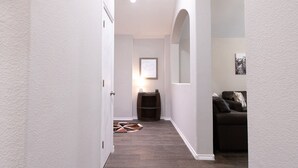 A hallway with a dark wood floor, white walls, a door on the left, a small table with a lamp, and a couch on the right. - Texas Roots: King Beds - 6 Minutes to Downtown McKinney- Sleeps 6 - Remodeled (McKinney)