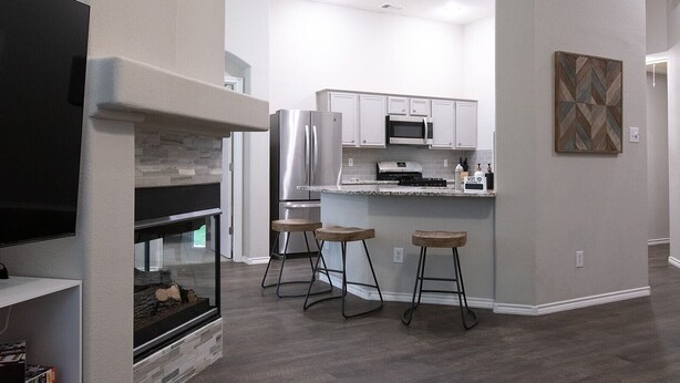 A modern kitchen with white cabinets, a stainless steel refrigerator, a microwave, and a built-in oven. There are three wooden stools and a fireplace with a stone surround. - Texas Roots: King Beds - 6 Minutes to Downtown McKinney- Sleeps 6 - Remodeled (McKinney)