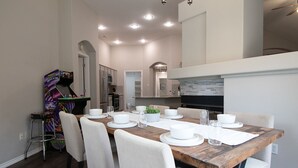 A dining area with a wooden table set for four, white chairs, and a kitchen in the background. - Texas Roots: King Beds - 6 Minutes to Downtown McKinney- Sleeps 6 - Remodeled (McKinney)
