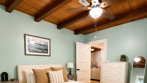 Une chambre à coucher comprenant un lit, des tables de chevet, une commode, un miroir, un ventilateur de plafond et une porte donnant sur une salle de bain.