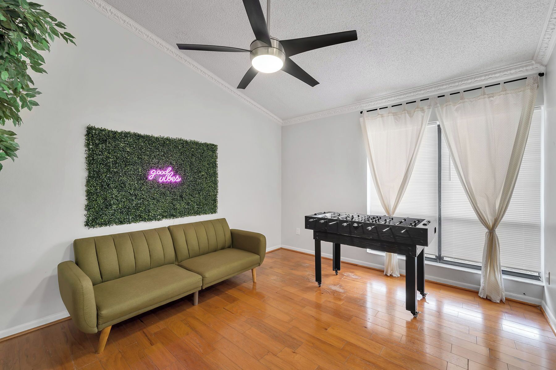 A room with a green sofa, a foosball table, a ceiling fan, and a wall-mounted green foliage decoration with a neon sign.