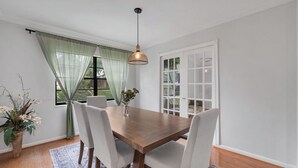 Comedor con mesa de madera, sillas blancas, lámpara colgante y vistas a través de una ventana con cortinas traslúcidas.