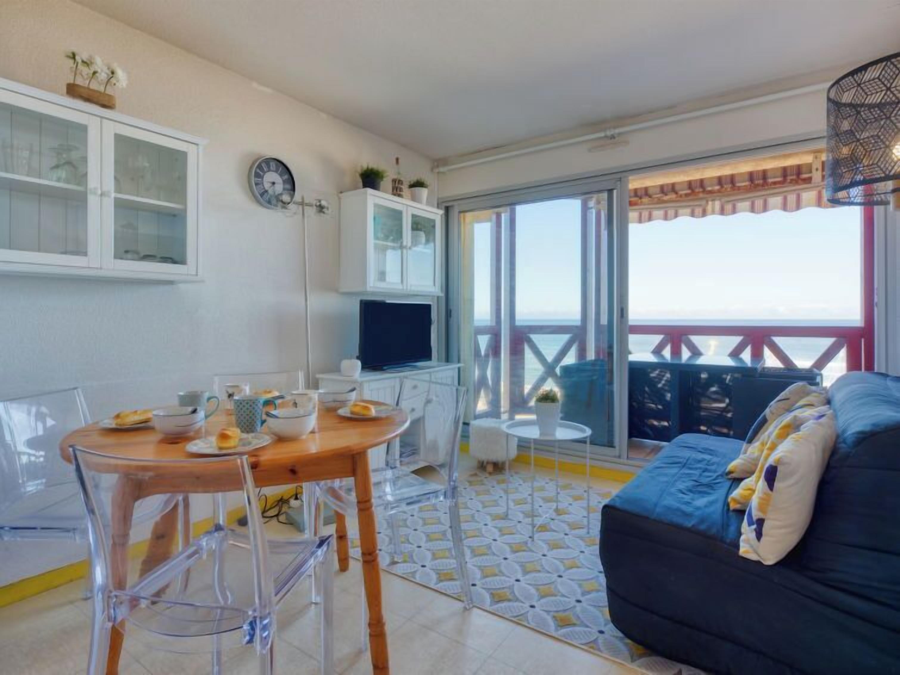 A modern living room with a dining area, a TV, and a balcony with a view of the sea.