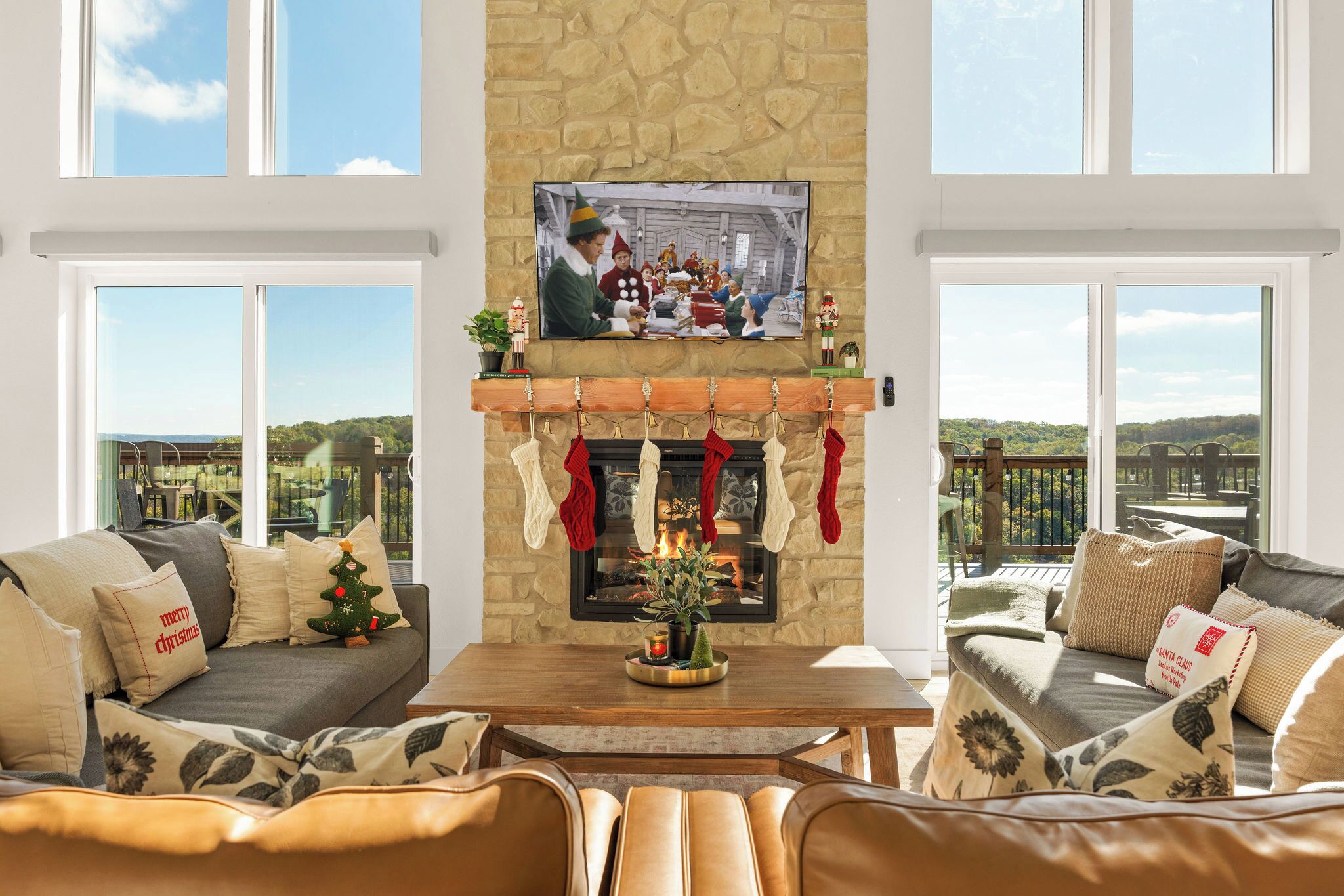 A cozy living room with a stone fireplace, a flat-screen TV mounted above it, and a scenic view through large windows.