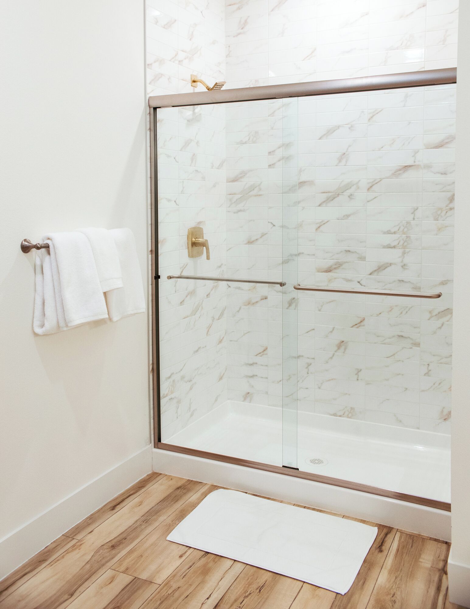 A glass-enclosed shower with marble tiles, a white towel on a hook, and a wooden floor.