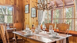 Een houten eetkamer met een grote tafel gedekt voor zes personen, afbeeldingen in lijsten aan de wanden en een kroonluchter met gewei-achtige takken.