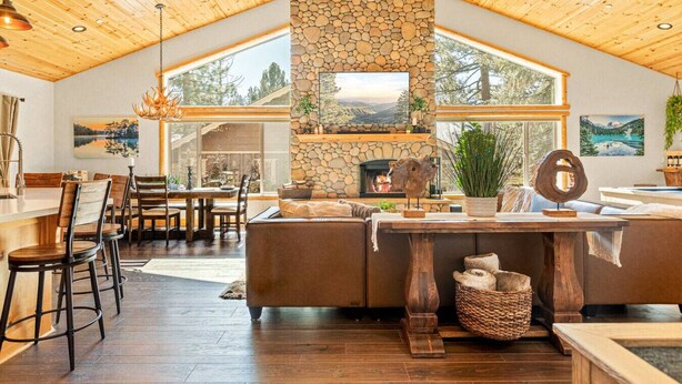 A spacious living area with a stone fireplace, wooden ceiling, and large windows offering views of the outdoors. - Stunning, like-new, 4-bedroom log-style cabin home. (Big Bear Lake)
