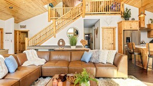 A spacious living room with a wooden staircase, a large leather sectional sofa, a dining area, and a kitchen with wooden cabinets. - Stunning, like-new, 4-bedroom log-style cabin home. (Big Bear Lake)