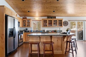 A kitchen with wooden cabinets, a stainless steel refrigerator, a large island with stools, and a view to the outside. - Fallen Leaf + A/C + Big Deck + BBQ + Games (Big Bear Lake)