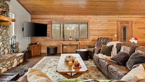 Interior acogedor de una cabaña con chimenea de piedra, un televisor de pantalla plana, un sofá de cuero y una mesa de comedor con una bandeja de comida y bebidas.