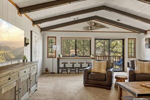 A spacious living area with a high ceiling, large windows, a wooden dining table, and a cozy seating arrangement.