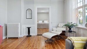 Ein Zimmer mit Holzboden, einem weißen Heizkörper, einem runden Tisch, einem weißen Stuhl, einem Bett mit weißer Bettwäsche und einem Fenster mit Vorhängen.