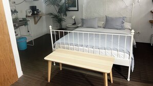 Un dormitorio con una cama de estructura metálica blanca, una mesita de noche, una lámpara, una planta en maceta y un cubo azul en el suelo.