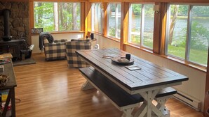A spacious room with a large wooden table, a stone fireplace, and large windows offering a view of a lake. - Cozy Lakeside Year Round Retreat ☀️🛶 🎣 🏊 🍁 🎃 🍎 🎿🏂 ❄️  & 🐾 are welcome 😊 (Hillsboro)