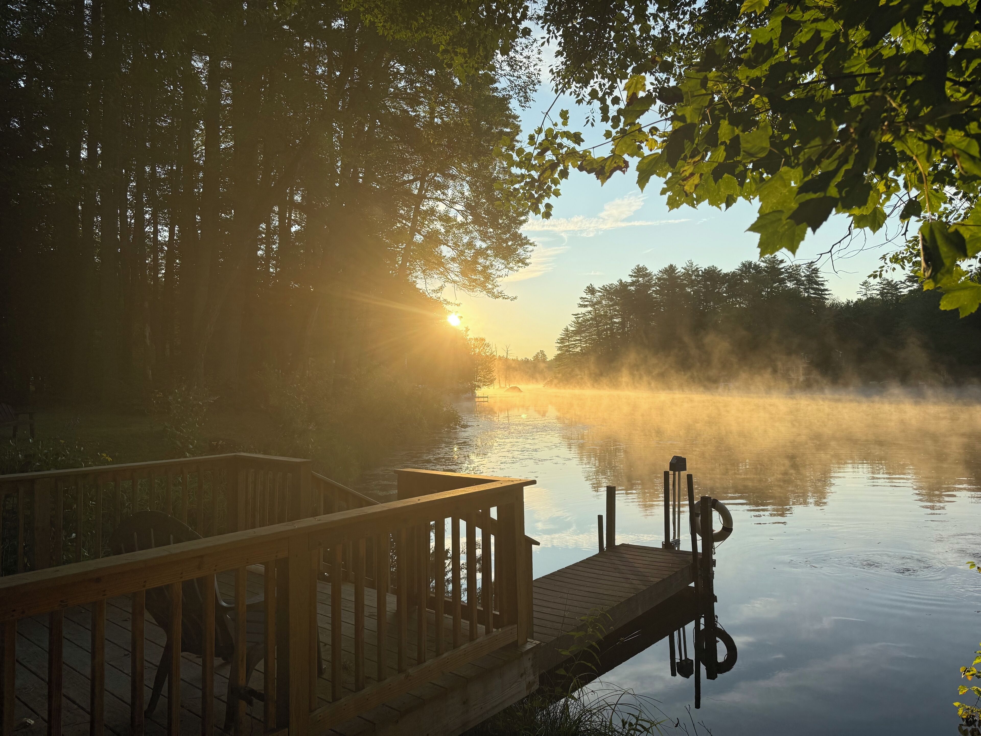Ein hölzerner Steg führt in einen nebligen See, im Hintergrund ein Sonnenaufgang.