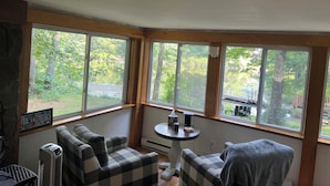 A cozy room with a checkered sofa, a small round table with a lamp, and large windows offering a view of a wooded area. - Cozy Lakeside Year Round Retreat ☀️🛶 🎣 🏊 🍁 🎃 🍎 🎿🏂 ❄️  & 🐾 are welcome 😊 (Hillsboro)
