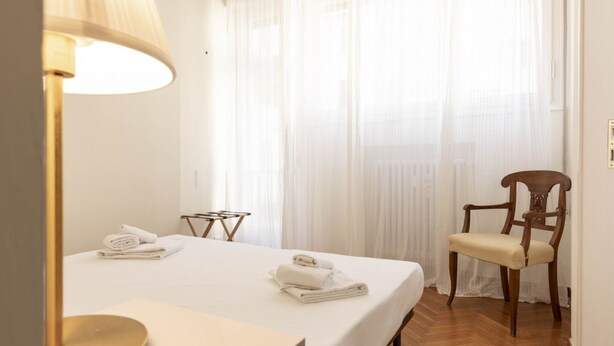 Ein Hotelzimmer mit Bett, Stuhl, Lampe und einem Fenster mit transparenten Vorhängen.