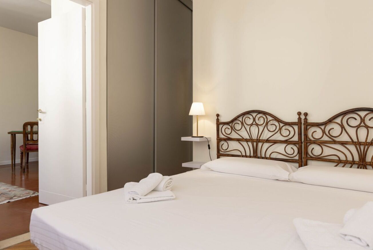 A neatly made bed with white linens and a decorative headboard, a bedside lamp, and a chair in the background.