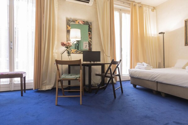 Une chambre d’hôtel comprenant un lit, un bureau avec un ordinateur, une chaise, une petite table et une fenêtre avec des rideaux.