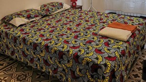 Quarto com colcha estampada, luminária vermelha, cabideiro e janela com cortina translúcida.