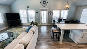 A spacious living room with a sofa, a dining area with a table and chairs, a kitchen island, and a large window with blinds. - Private Beachfront House on Bolivar Peninsula – Clean, Quiet & Steps to Sand (Bolivar Peninsula)